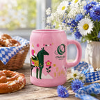 Rosa Oktoberfest-Bierkrug aus Steingut mit München-Motiv, Brezen und Blumen auf einem hellen Tisch