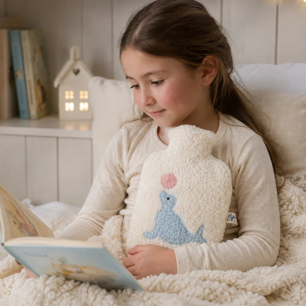 Kind liest im Bett und wärmt sich mit der EFIE Kinder-Wärmflasche mit Seehundmotiv.