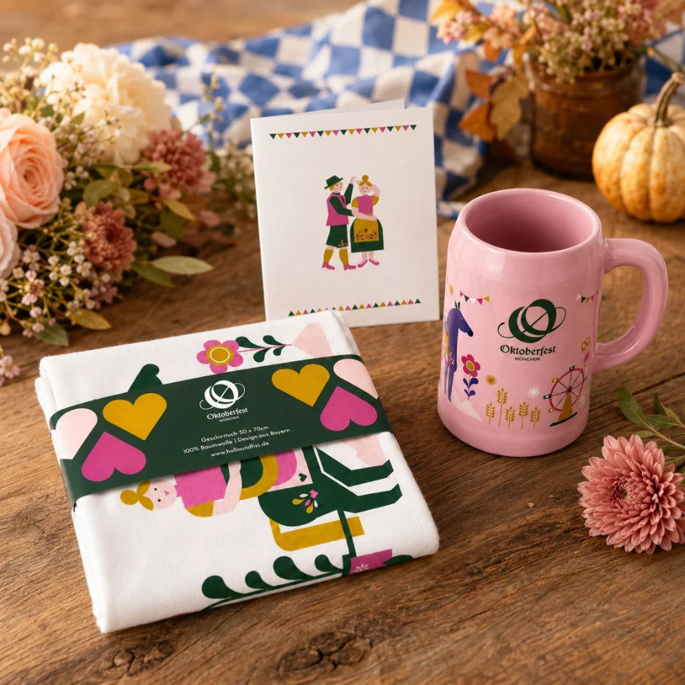 Oktoberfest-Geschenkbundle für sie mit rosa Bierkrug, Geschirrtuch und Postkarte auf herbstlich dekoriertem Holztisch.