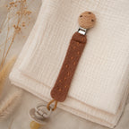 Braune Schnullerkette aus Bio-Baumwolle mit Holzclip und befestigtem Schnuller auf Musselin