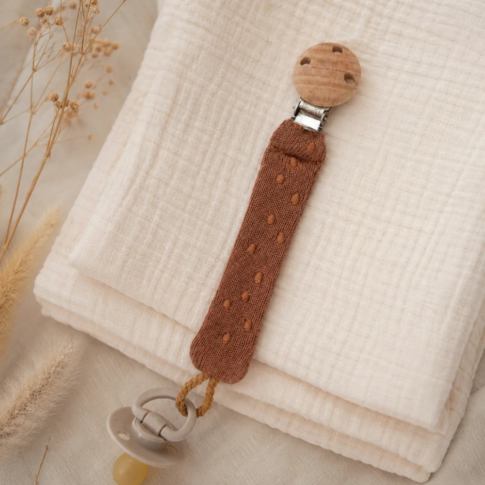 Braune Schnullerkette aus Bio-Baumwolle mit Holzclip und befestigtem Schnuller auf Musselin